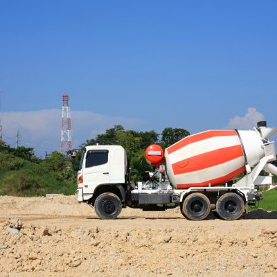 White and red concrete mixer truck parked on a construction site with a clear blue sky and lush greenery in the background.